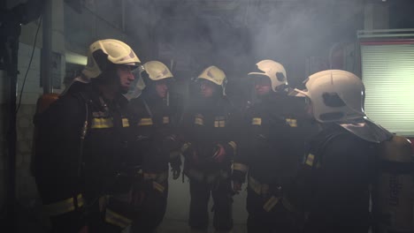 group of five firefighters wearing full equipment in smokey area. relaxed chatting after work well done. fire trucks in the background. name tag translation is fireman and model's last name.