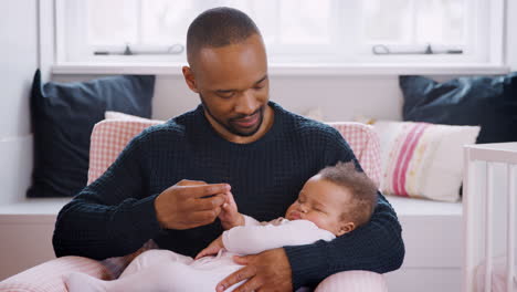 new father sitting in chair holding hands with sleeping baby girl in nursery at home