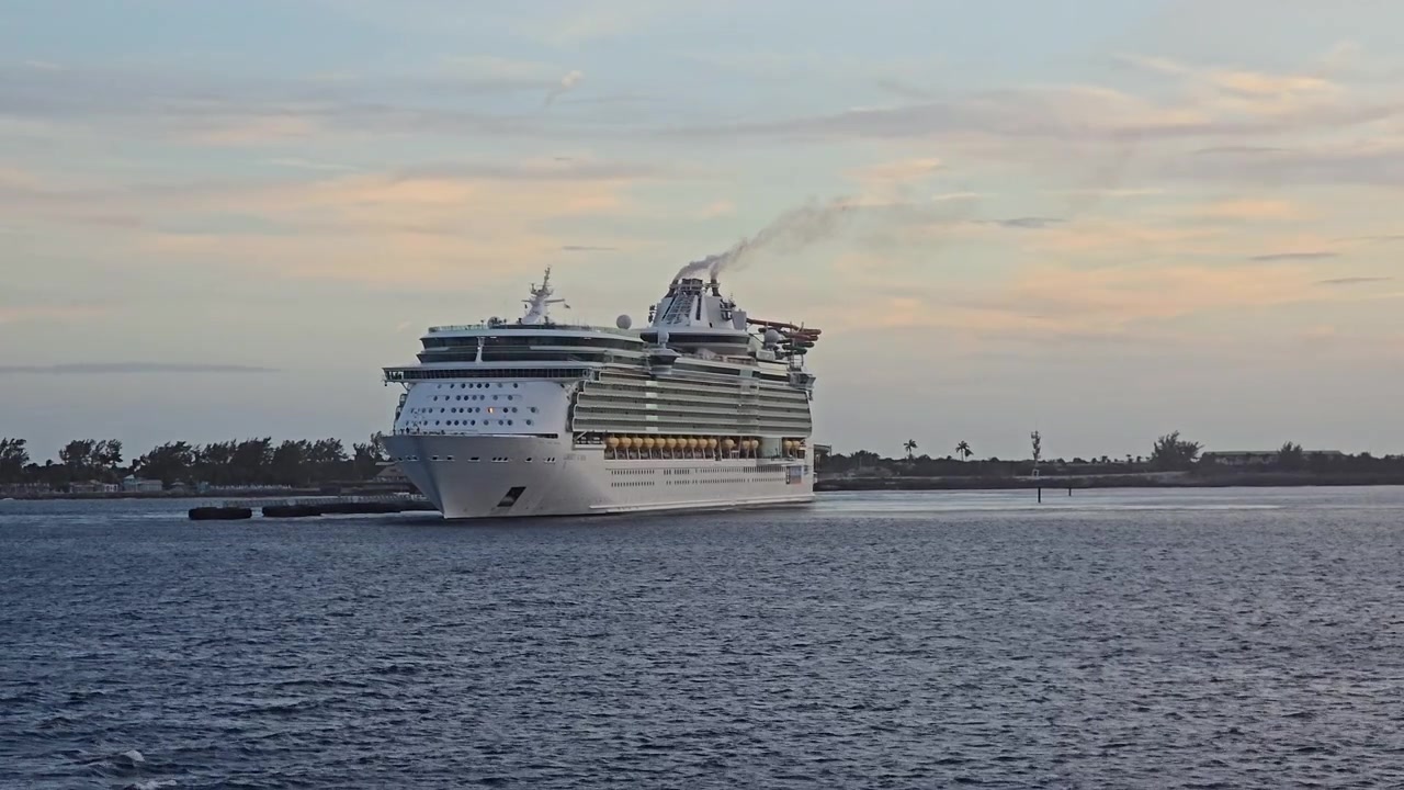 Et stort luksuspassagerkrydstogtskib sejler på roligt havvand nær en kystlinje i skumringen. Røg stiger op fra skorstenen, mens fartøjet navigerer mod sin destination under en skyet himmel.