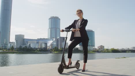 Young-Elegant-Businesswoman-Standing-On-Electric-Scooter-By-Riverside-Against-Modern-Buildings-On-Sunny-Day