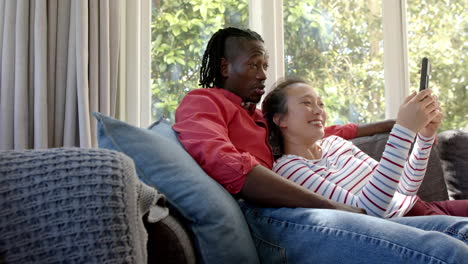 happy diverse couple relaxing on couch using tablet in living room, copy space, slow motion