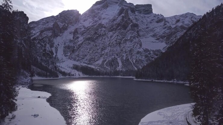 Superficie di un lago ghiacciato incastonata tra montagne rocciose innevate e fitte foreste di conifere durante l'inverno. Lago di Braies. Paesaggio selvaggio e freddo con terreno ghiacciato e cime maestose