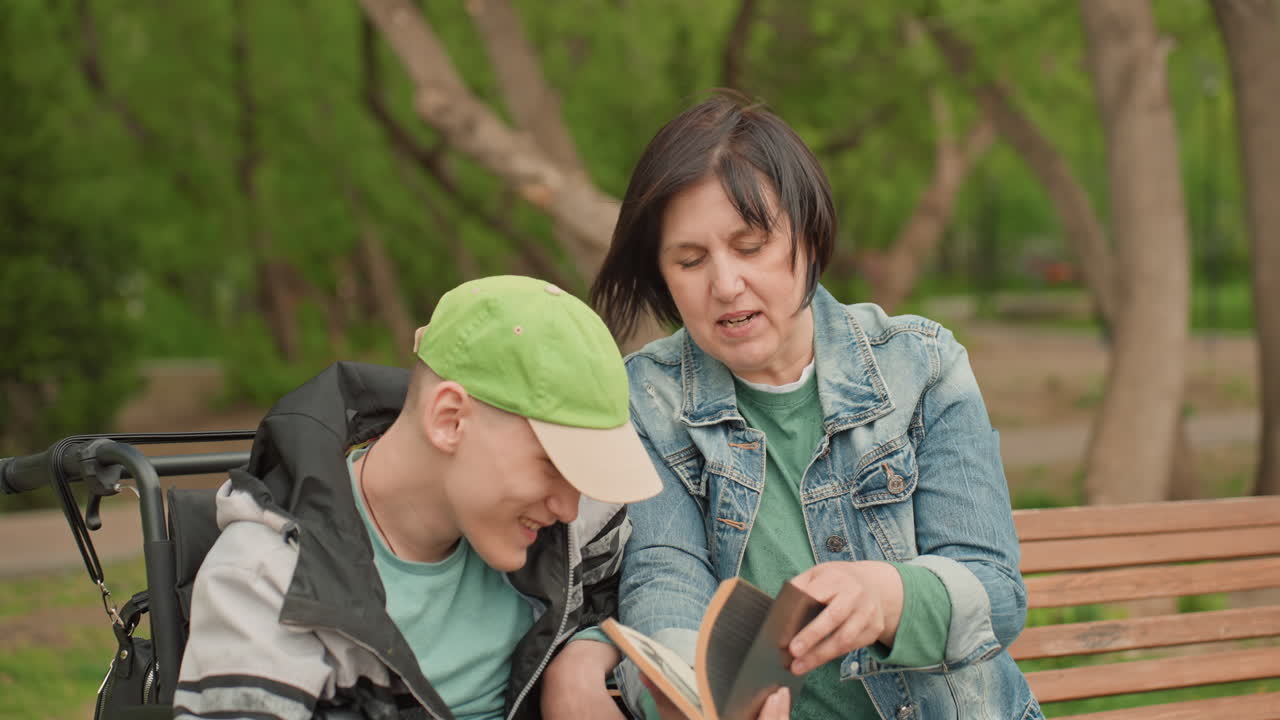 White Caregiver Reading Aloud To White Teen In Wheelchair On Park Bench With Open Book And Attentive Gestures Inclusive Education Moment, Denim Jacket, Green Cap, Supportive Conversation And Calm