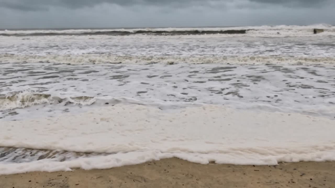 Close-up view of foamy waves gently washing over a sandy beach under overcast skies.
