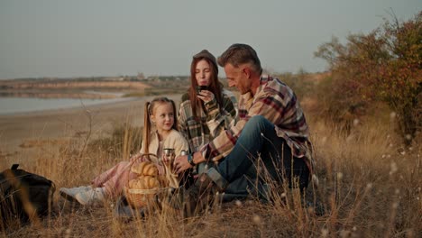 A-happy-brunette-man-with-gray-hair-in-a-checkered-shirt-sits-on-the-ground-on-a-special-mat-during-a-picnic-and-drinks-tea-from-a-thermos-with-his-wife-and-little-daughter-during-a-picnic-outside-the-city-near-a-pond-in-summer