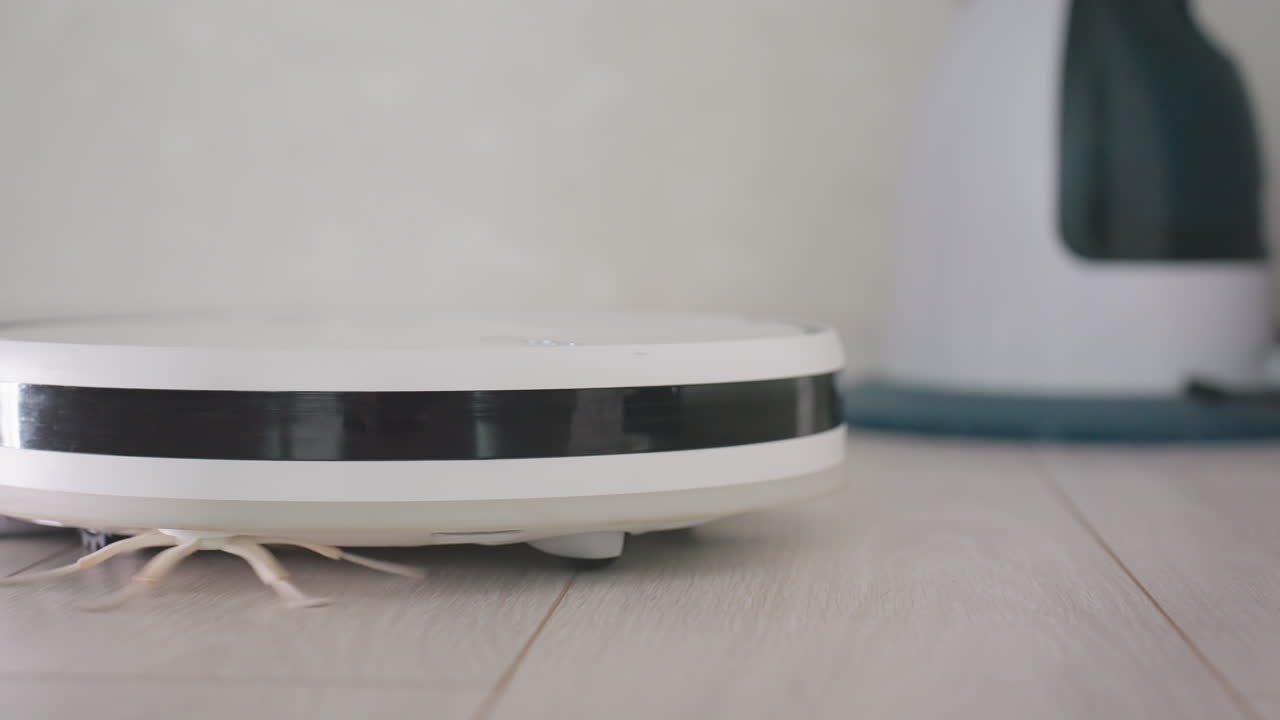 Robot vacuum cleaner rotates while moving across wooden floor, capturing detail of smart cleaning device in action with blurred view of another household gadget close to wall in modern interior