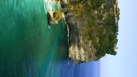 Vertical-Reveal-shot-of-the-cliffs-at-Uluwatu,-near-the-Uluwatu-temple-with-brilliant-green-waves-crashing-against-the-greenery-covered-cliff-walls