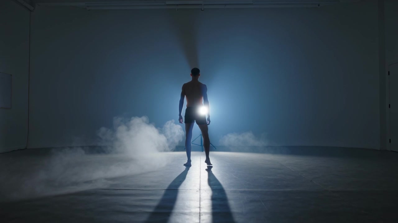 Homem sem camisa de shorts movendo-se no estúdio, luz azul se intensificando, névoa se espalhando.
