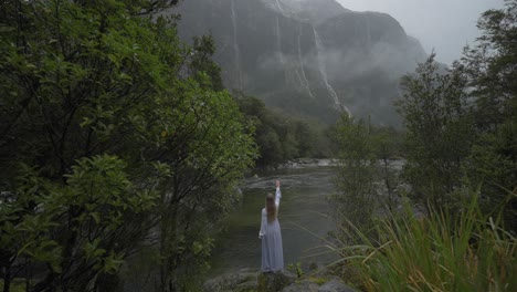 Dama-Con-Vestido-Largo-Parada-En-La-Orilla-Rocosa-Del-Río-Con-Cascadas-Distantes,-Escena-Mágica