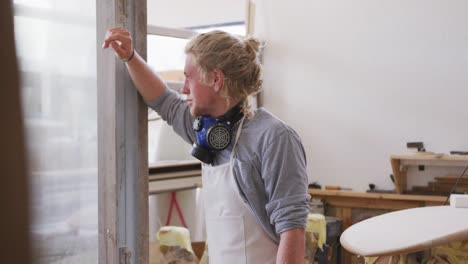 caucasian male surfboard maker wearing a face mask and standing in his studio