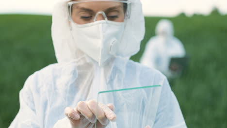 rear view of researcher woman in protective suit and goggles tapping on glass in the green field