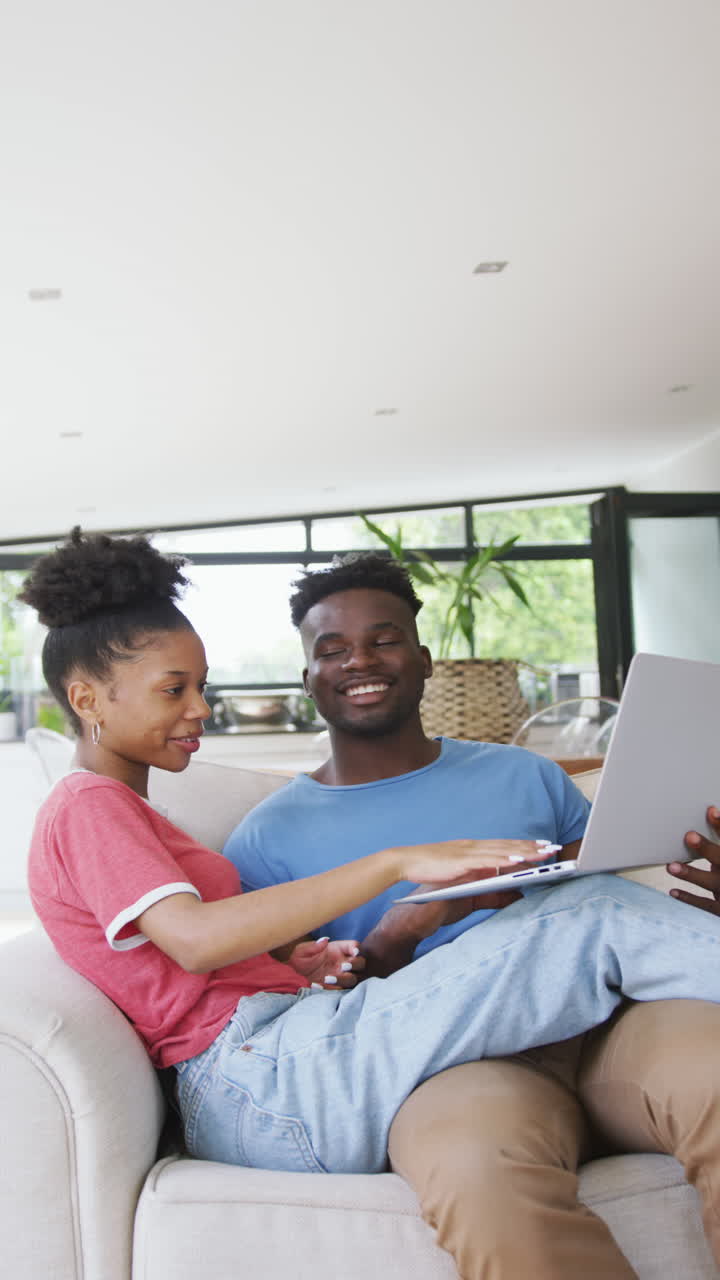 Vertical video of happy african american couple using laptop at home