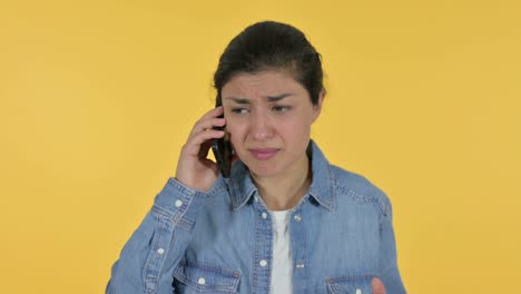 angry young indian woman talking on smartphone, yellow background