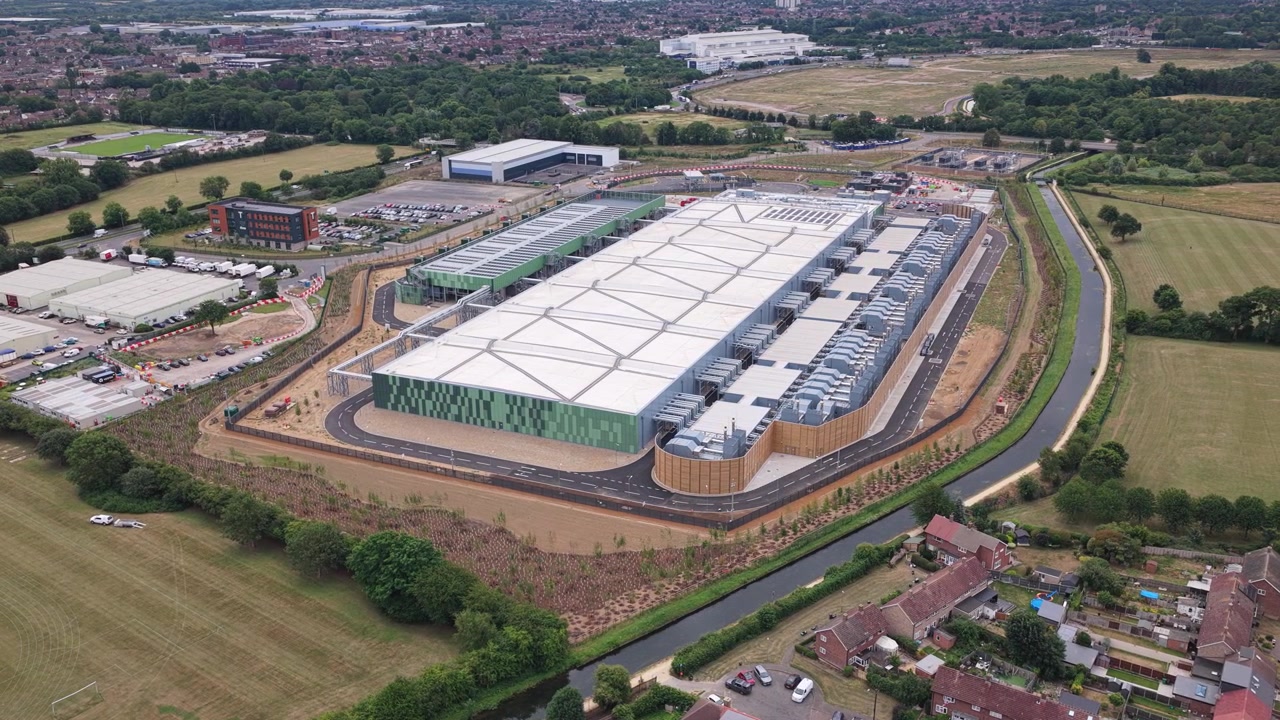 Grand bâtiment de centre de données industriel vu du ciel dans une zone rurale avec des champs d'herbe et des routes environnants sous une lumière du jour éclatante pendant la saison estivale.