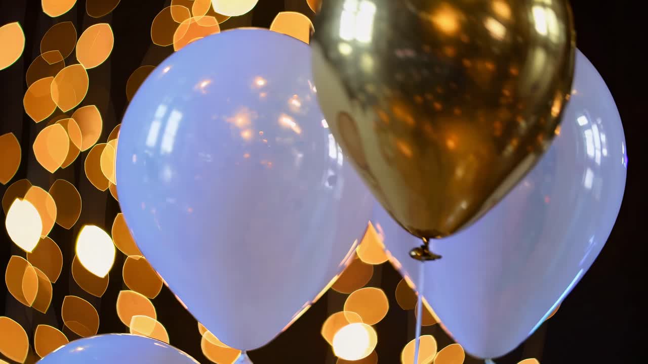 A festive video scene with a close-up angle of white and gold balloons against a backdrop of warm