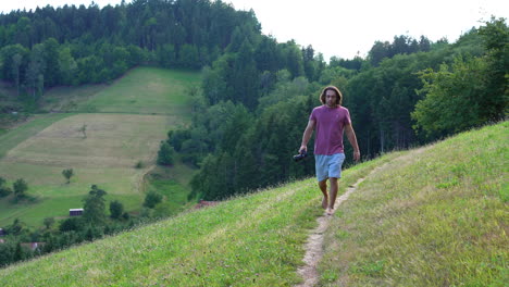 Young-man-in-red-T-Shirt-and-long-hair-walks-barefoot-in-slowmotion-towards-the-camera-on-a-small-path-on-a-hill-on-a-sunny-summer-Day