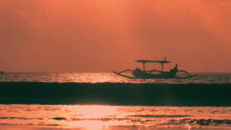 Wave-Rolling-Towards-Sandy-Beach-And-Breaking-with-Splashes-Backlit-with-Sunset-Sunlight,-Outrigger-Old-Boat-in-Silhouette-Anchored-Floating