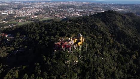 drone shot of historic pena palace castle illuminated by sunlight standing a top of a hill in sintra mountains above town of sintra, portugal