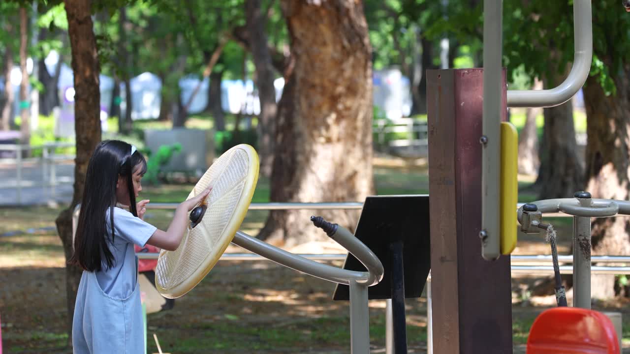 A young Asian girl spins a yellow exercise wheel at an outdoor public park gym