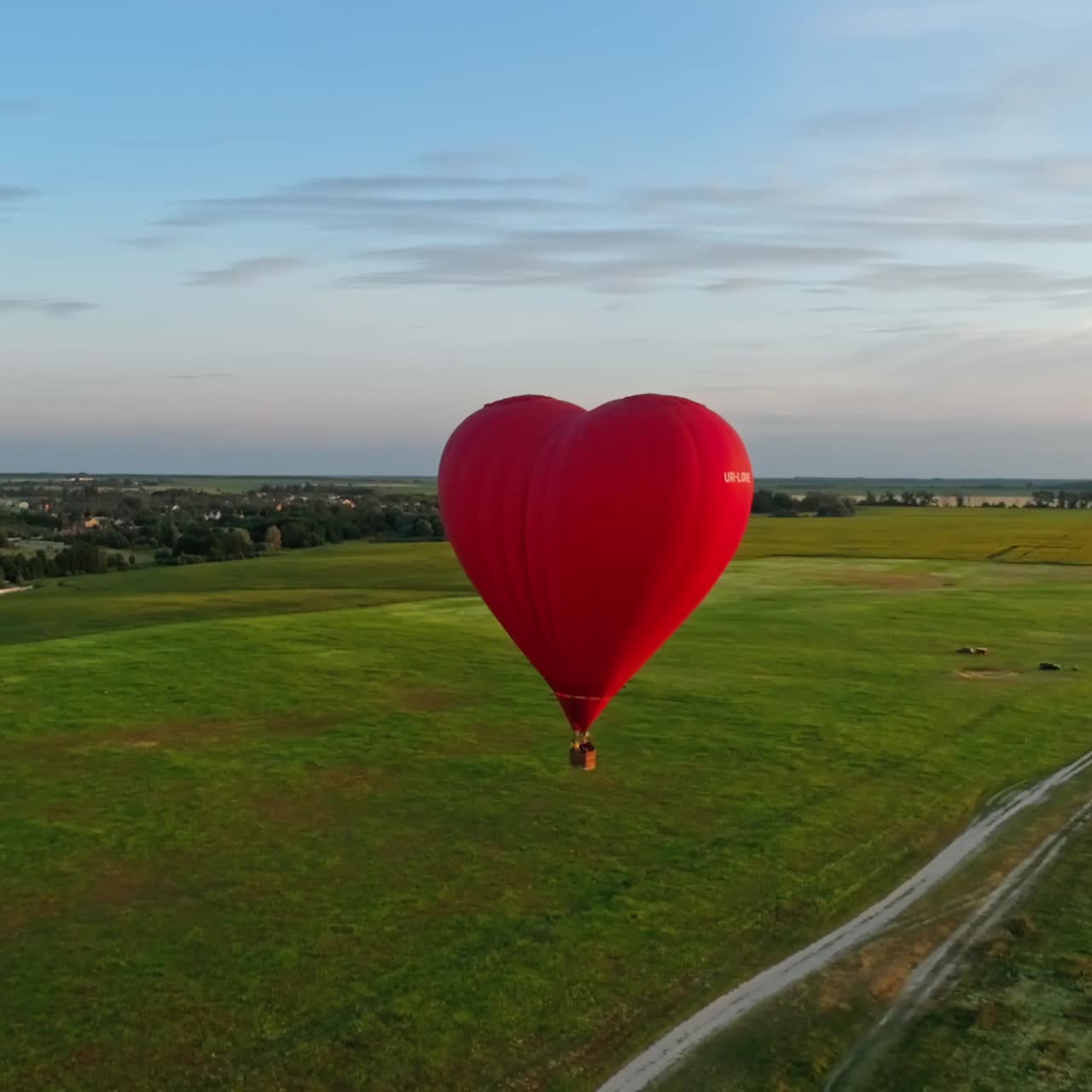 Heart shape aerostat against the setting sun. Red hot air balloon with basket flying over the green fields at sunset. Romantic airship in summer.