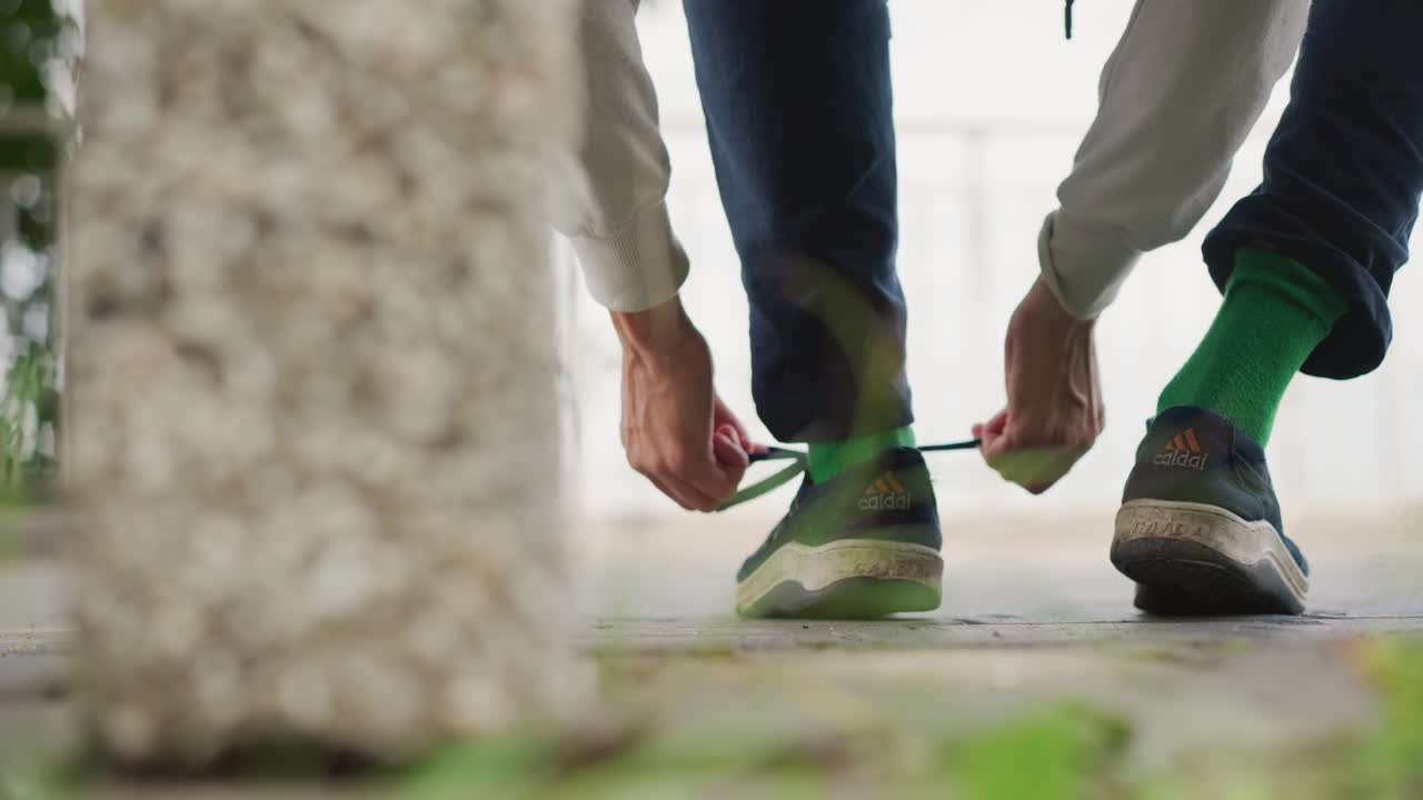 달리는 사람이 젖은 아침, 콘크리트 기둥 근처에서 달리기를 준비하며 포장 도로에서 초록색 끈을 묶고 있어요. 클로즈업한 손과 발, 이끼와 웅덩이 디테일, 결연한 분위기와 미묘한 움직임 텍스처가 돋보입니다.