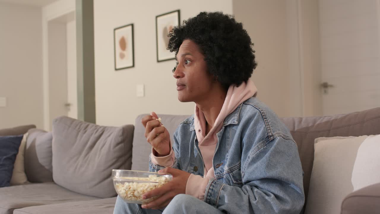 Afro-americano de meia-idade reagindo a um evento fora da tela enquanto come pipoca no sofá, espaço para texto