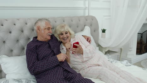 Senior-elderly-couple-wearing-pyjamas-lying-on-bed-looking-on-mobile-phone-making-online-shopping