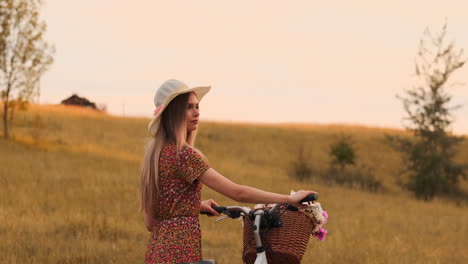 Back-kind-of-Happy-blonde-girl-in-dress-and-hat-turns-around-and-smiling-cheerfully-looks-at-the-camera-and-flirts-strolling-around-the-field-in-summer-with-bike-and-flowers.