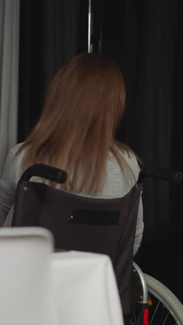 young woman moves to window in wheelchair adjusting hair with hand. lonely housewife with spinal cord injury sits in kitchen staying late closeup