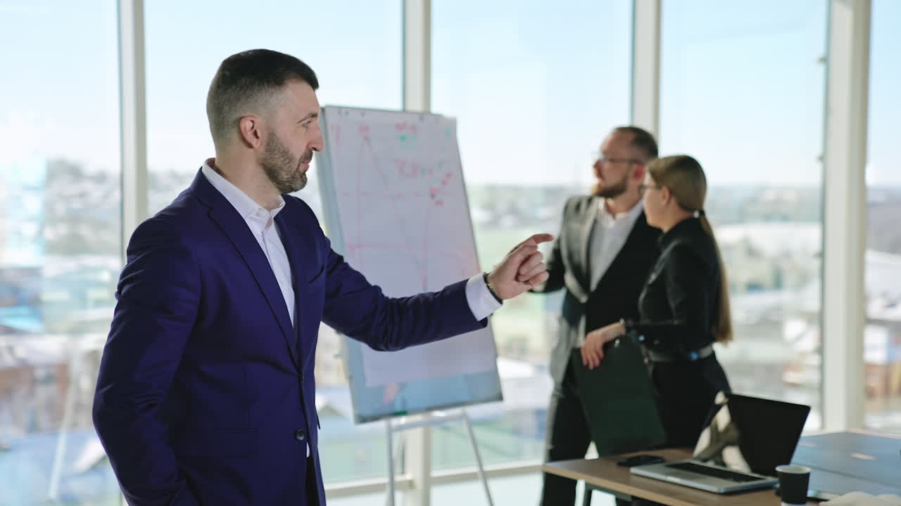 Bearded businessman smiling and talking to the camera. Successful professional standing in the office. Two office people at the backdrop.