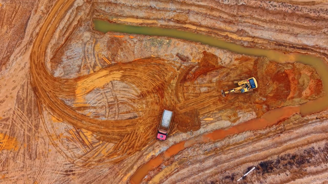 Top down view of dump truck and excavator at mining site. Overhead drone capture of two machines on orange clay terrain with muddy water channels.