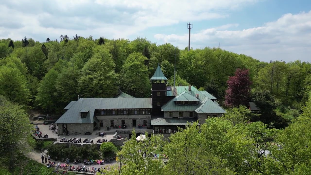 Dronové záběry horské chaty Szyndzielnia obklopené bujnými lesy, turistickými stezkami a panoramatickými beskydskými krajinami za slunečného letního dne s návštěvníky relaxujícími venku