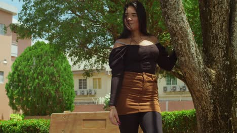 a young hispanic girl stands next to a tree at the park in a mini skirt