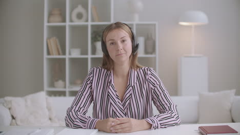 young friendly woman is listening interlocutor by video chat by headphones and nodding looking at camera