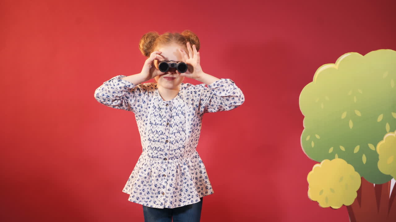 little girl in print blouse are looking through binoculars around herself on the background of red studio and scenery of trees