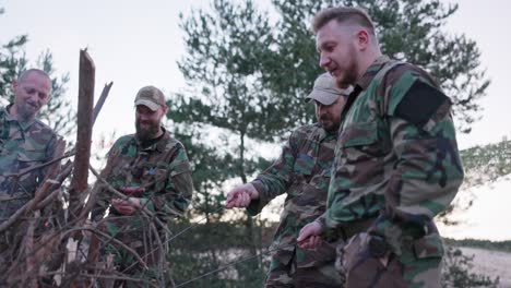 conversación de soldados en ropa de camuflaje, están discutiendo por la noche junto a la fogata común fritando salchichas para la cena, los hombres están descansando después del trabajo, servicio, ejercicios de campo