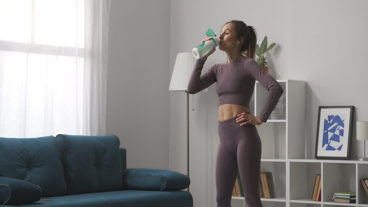 giovane donna atletica stanca sta bevendo acqua dopo l'allenamento di fitness a casa ritratto medio nel soggiorno rilassarsi e riposare stile di vita sano