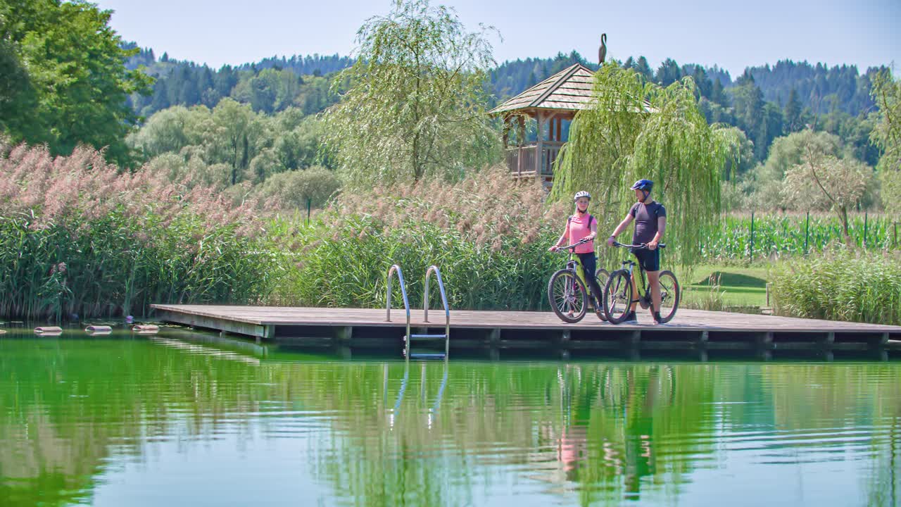 Cyclist Couple Relaxes On A Pier By A Lake. Radlje ob Dravi, Slovenia