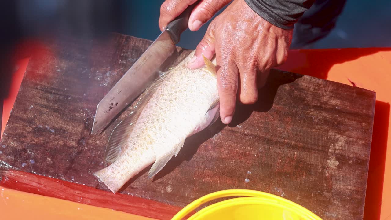 A person fillets a fish on a wooden board aboard a boat in Phuket, Thailand, under bright sunlight