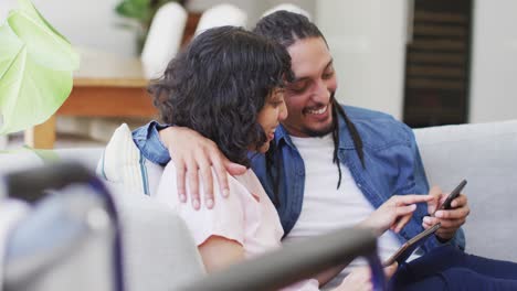 Happy-biracial-couple-in-living-room-using-laptop-and-talking,-with-her-wheelchair-in-foreground