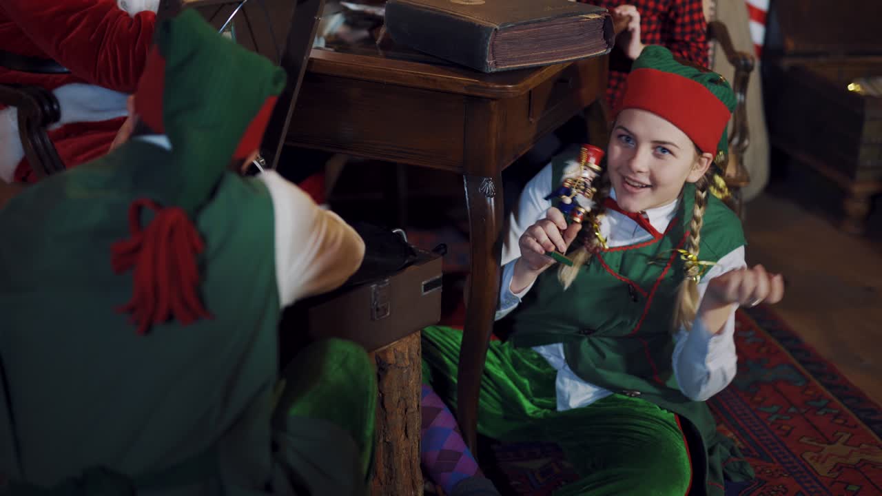 Fairy characters in green costumes play with a toy at Christmas. Happy female elf holding a toy soldier and shows it to another elf in the room.