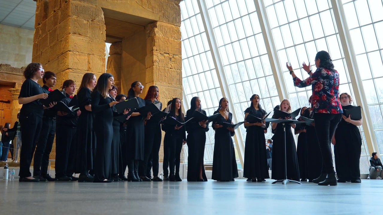 New York, USA, 18 marzo 2026: Coro giovanile che esegue musica vocale in un'ala di un museo storico. Giovani cantanti in nero che tengono cartelle si esibiscono con un direttore d'orchestra vicino a un antico monumento in pietra e una parete di vetro.