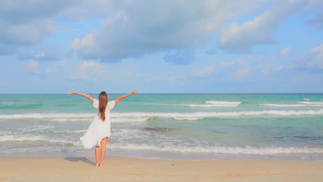 Zeitlupe-Einer-Frau,-Die-Im-Sommer-Barfuß-Am-Strand-In-Richtung-Meer-Geht-Und-Springt,-Dann-Hält-Sie-An-Und-Hebt-Die-Hände-Hoch,-Handgehaltene-Rückseitenansicht