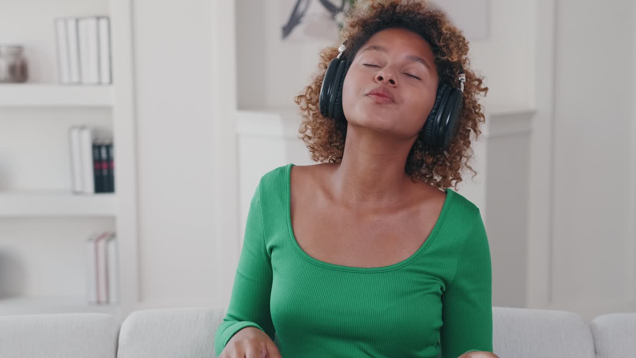 Jovem afro-americana relaxada de fones de ouvido dança sentada no sofá