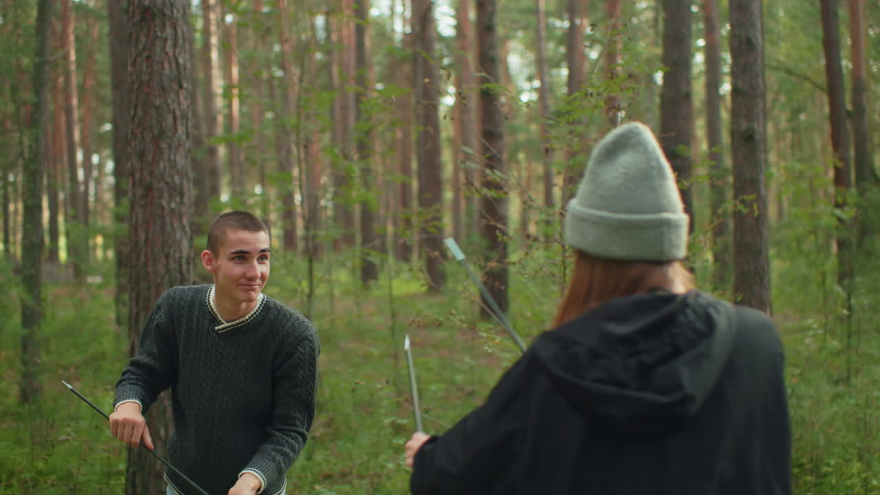Young man smiles joyfully while playing with friend in forest, holding tent poles during fun outdoor camping moment surrounded by trees and natural scenery in relaxed woodland setting