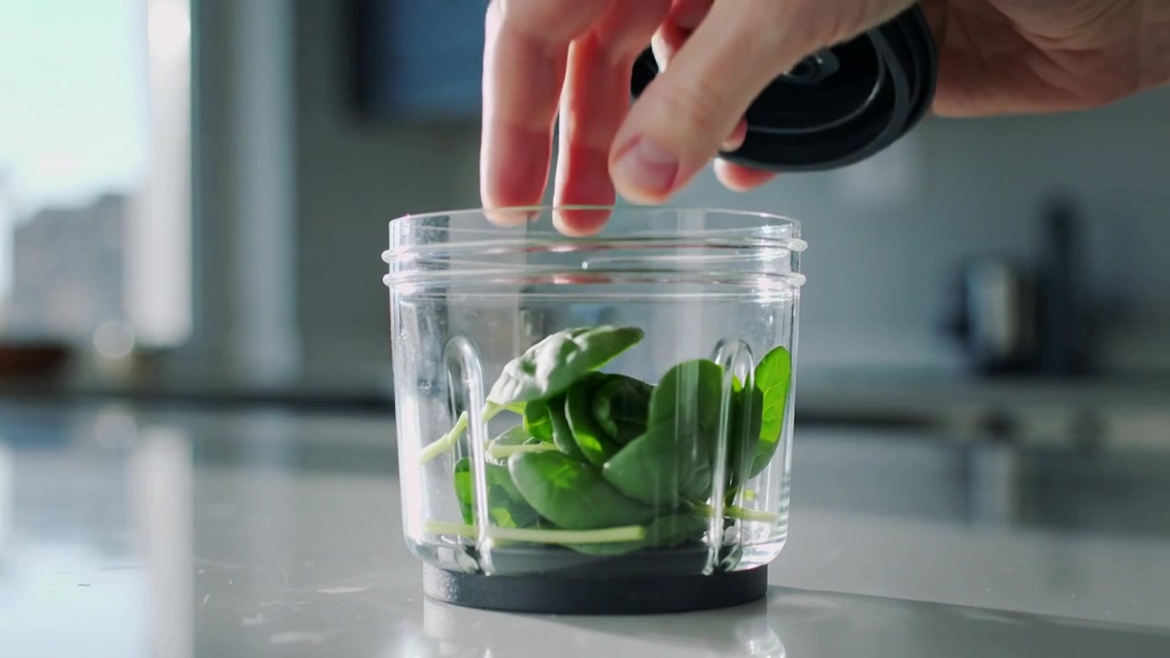 Hands lifting spinach, placing leaves in blender cup on kitchen counter locking blade lid to blend
