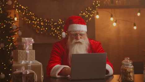 Real-Santa-Claus-using-new-technology-for-communication-with-children-receiving-mail-or-wish-list.-Cheerful-working-on-laptop-and-smiling-while-sitting-at-his-chair-with-fireplace-and-Christmas-Tree.-High-quality-4k-footage