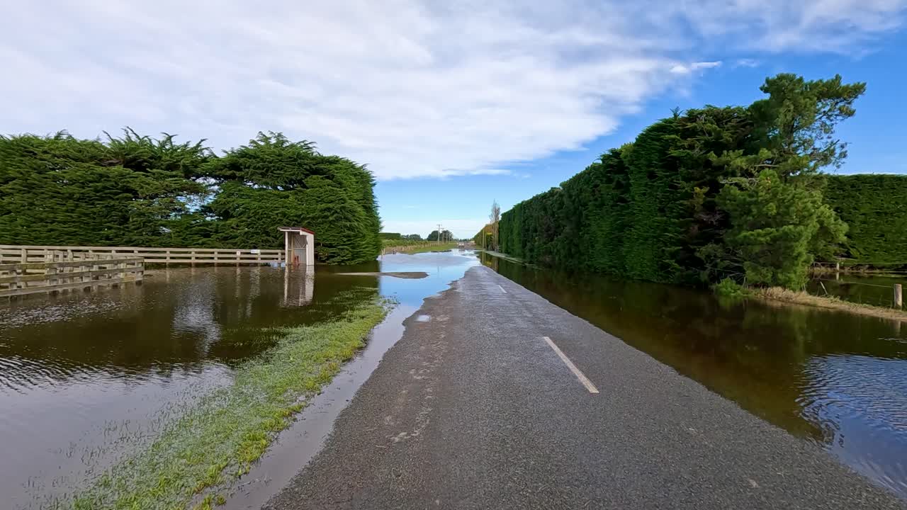 A vehicle travels along a rural road partially submerged by floodwater, bordered by hedges and farmland under bright daylight with steady camera movement