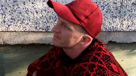 adult male wearing a black and red sweater and hat in the wind against the side of a house