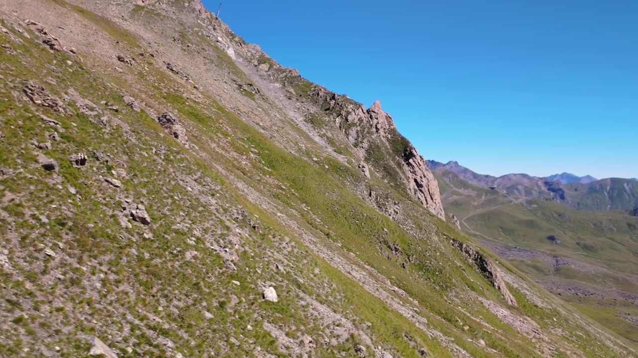 Discesa in seggiovia su un versante alpino soleggiato, erba verde, pendii rocciosi e una cresta montuosa distante sotto un cielo estivo limpido e azzurro